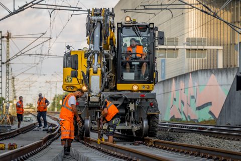 Bauarbeiter bei Gleisarbeiten mit Baumaschine in urbaner Umgebung.