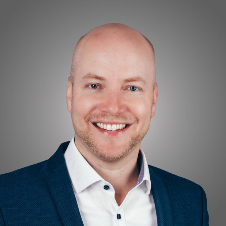 Mann in Businesskleidung lächelt vor grauem Hintergrund.