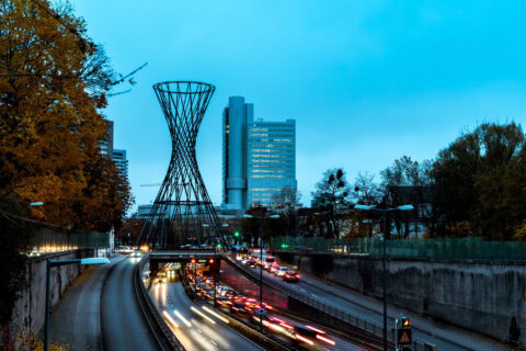 Verkehrsreiche Straße mit Brücke und moderner Stadt im Hintergrund.